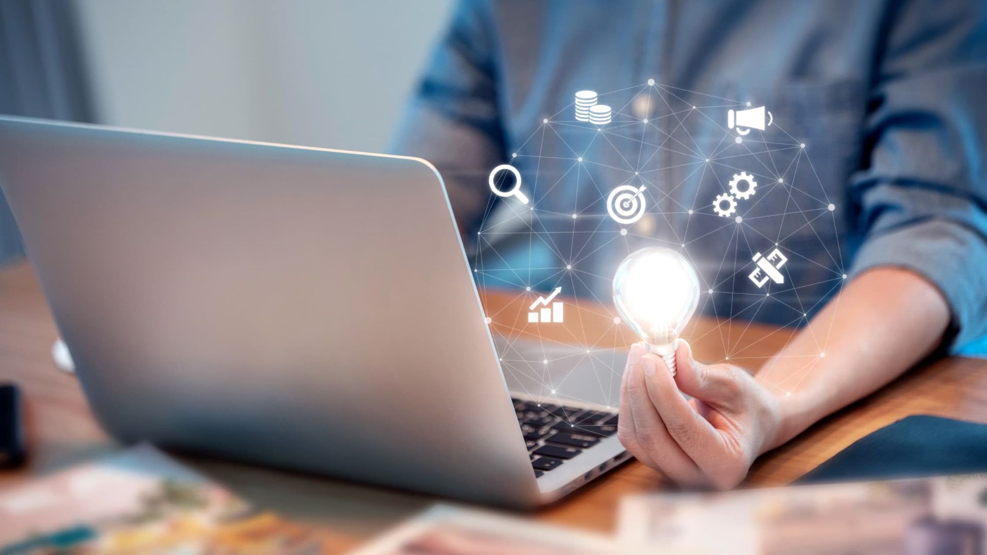 Une personne assise à un bureau avec un ordinateur portable, tenant une ampoule allumée entourée d'icônes numériques liées au commerce et à la technologie.