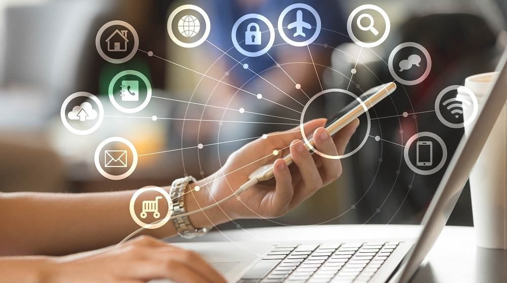 Une main tient un smartphone connecté à un réseau numérique, affichant diverses icônes de communication et de services au-dessus d'un ordinateur portable.
