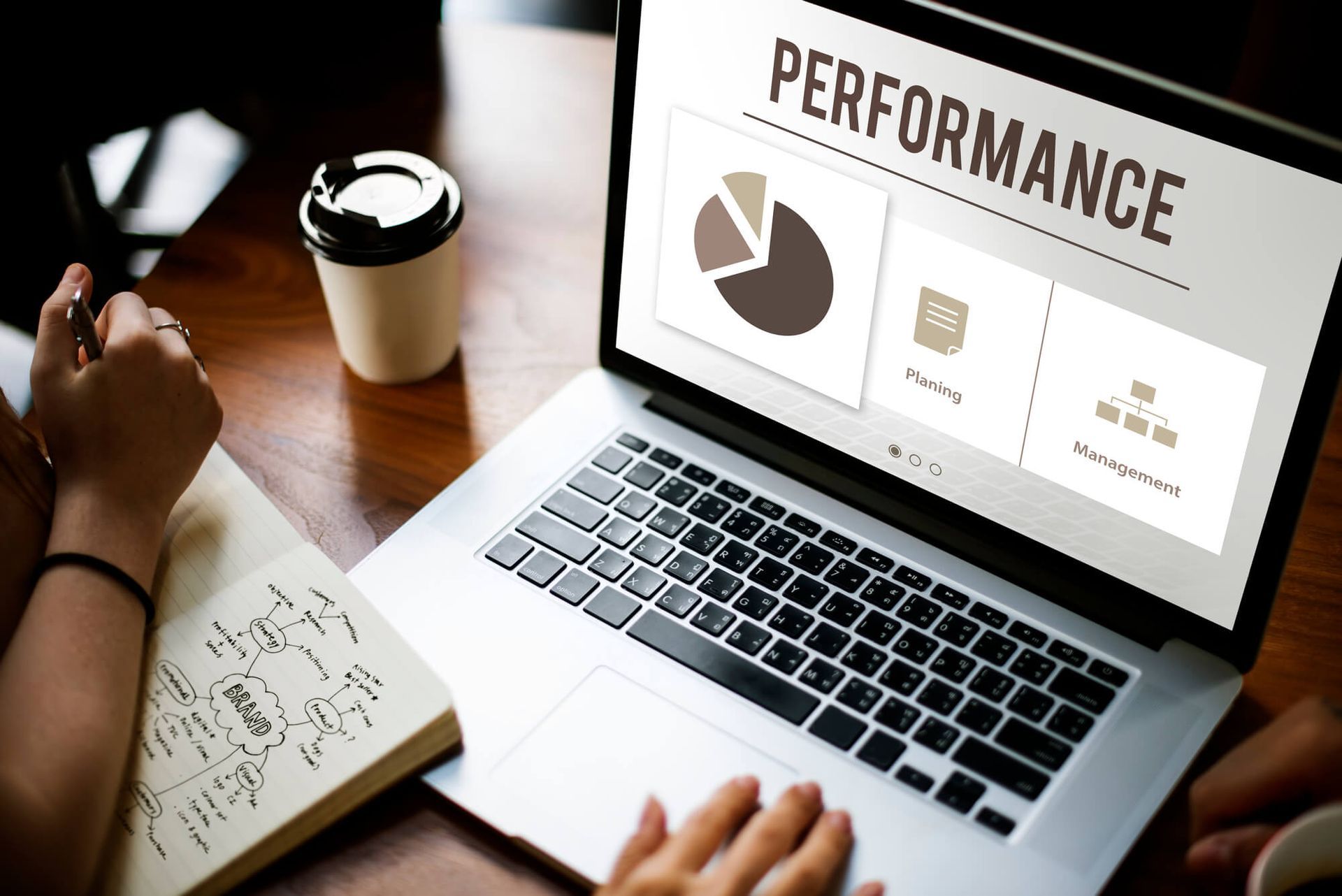 Une personne utilise un ordinateur portable affichant des graphiques et des diagrammes de performance à un bureau en bois, avec un carnet et une tasse de café.