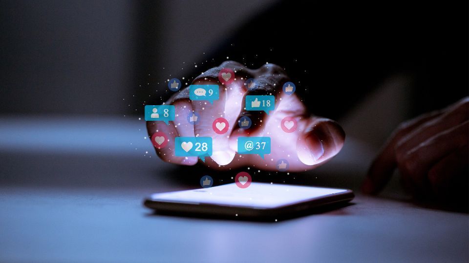 Une main plane au-dessus d'un smartphone lumineux, entouré d'icônes de notifications de réseaux sociaux flottantes dans un environnement sombre.