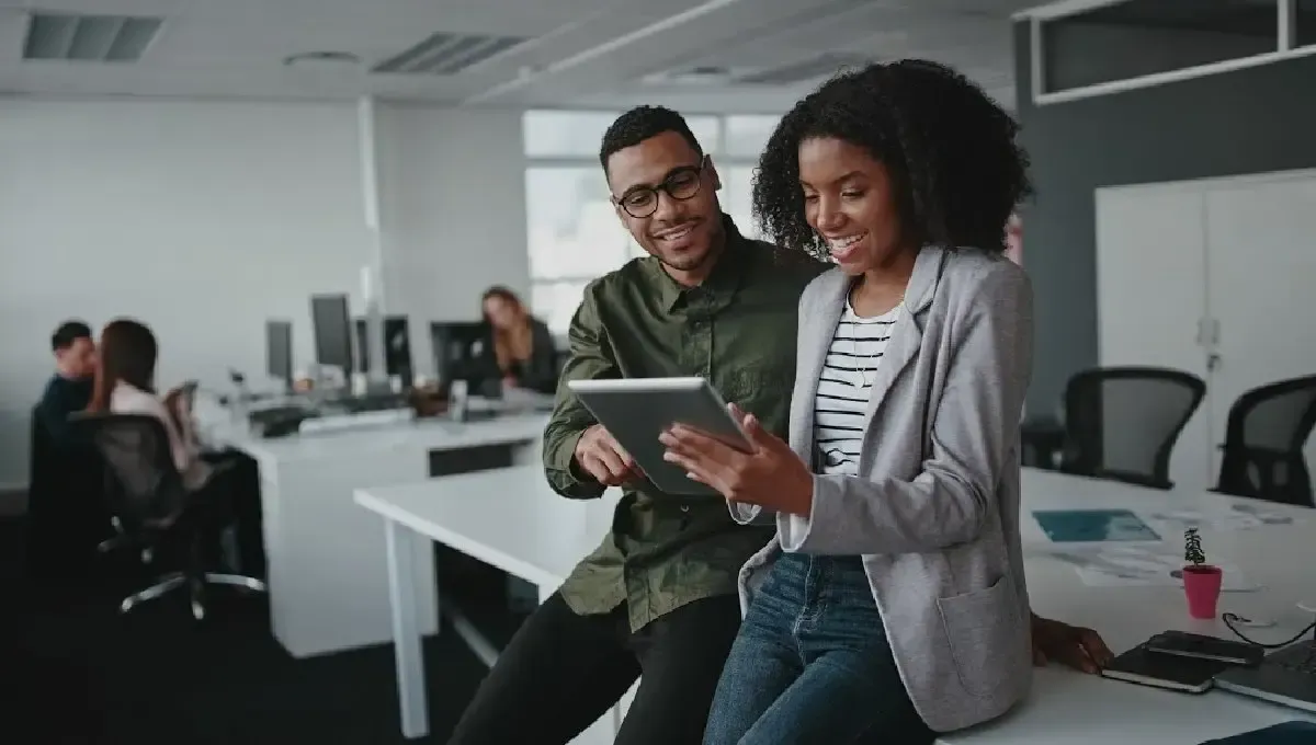 Deux collègues sourient en consultant ensemble une tablette dans un décor de bureau moderne et flou.