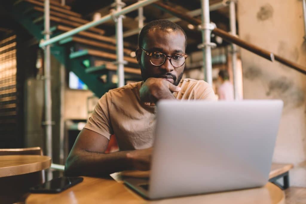 Une personne concentrée, portant des lunettes, pose son menton sur sa main tout en regardant un ordinateur portable ouvert à la table d'un café.