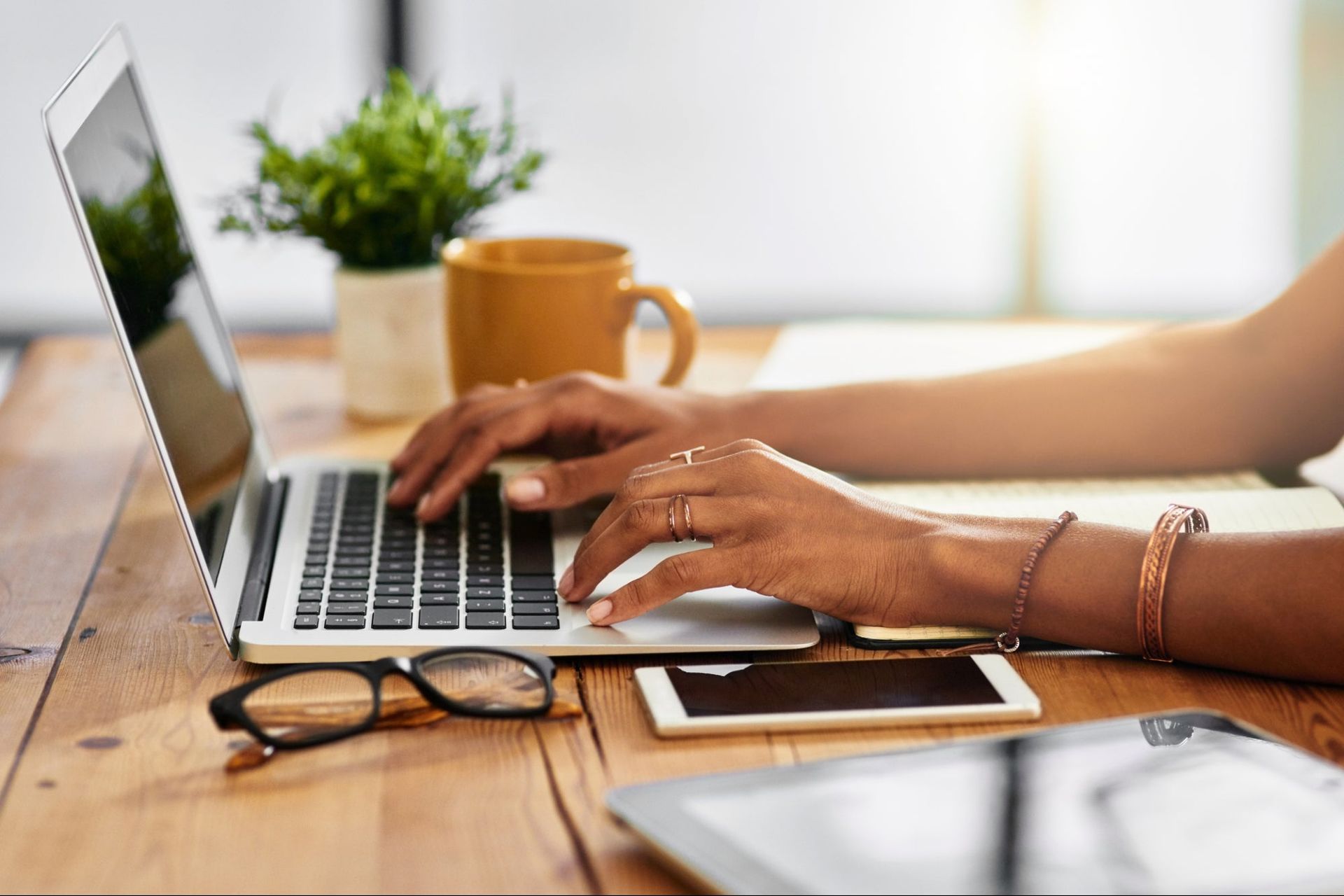 Des mains tapent sur un ordinateur portable argenté posé sur un bureau en bois, avec une plante en pot, une tasse à café, des lunettes et un téléphone à proximité.