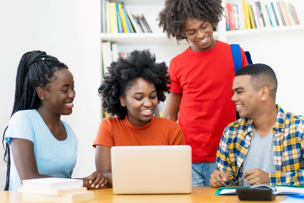 Quatre étudiants sourient en travaillant ensemble sur un ordinateur portable à une table de bibliothèque.