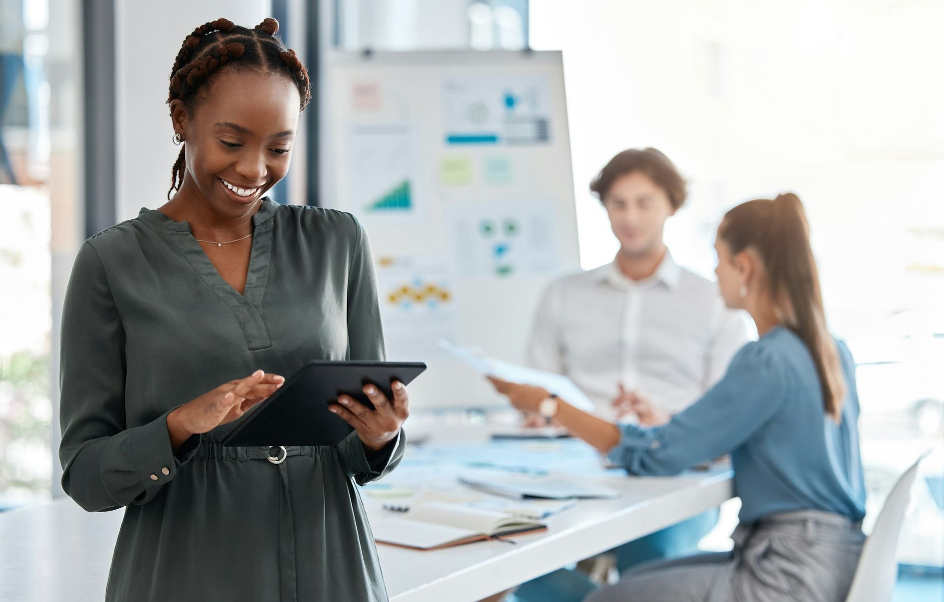 Dans un bureau lumineux, une femme souriante utilise une tablette numérique, tandis que ses collègues collaborent à une table en arrière-plan.