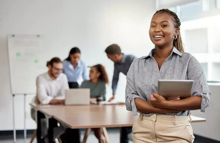 Une personne souriante tient une tablette dans un bureau, tandis que des collègues collaborent autour d'une table de conférence en arrière-plan.