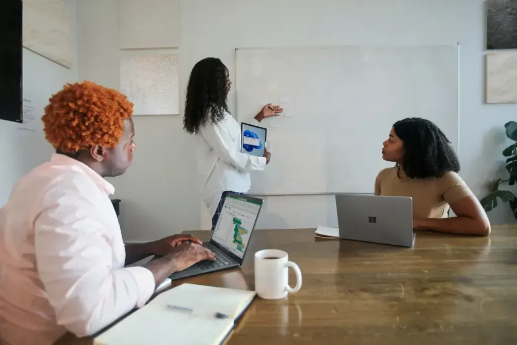 Trois personnes collaborent dans un bureau ; l’une d’elles désigne un tableau blanc vierge tandis que les deux autres utilisent des ordinateurs portables.