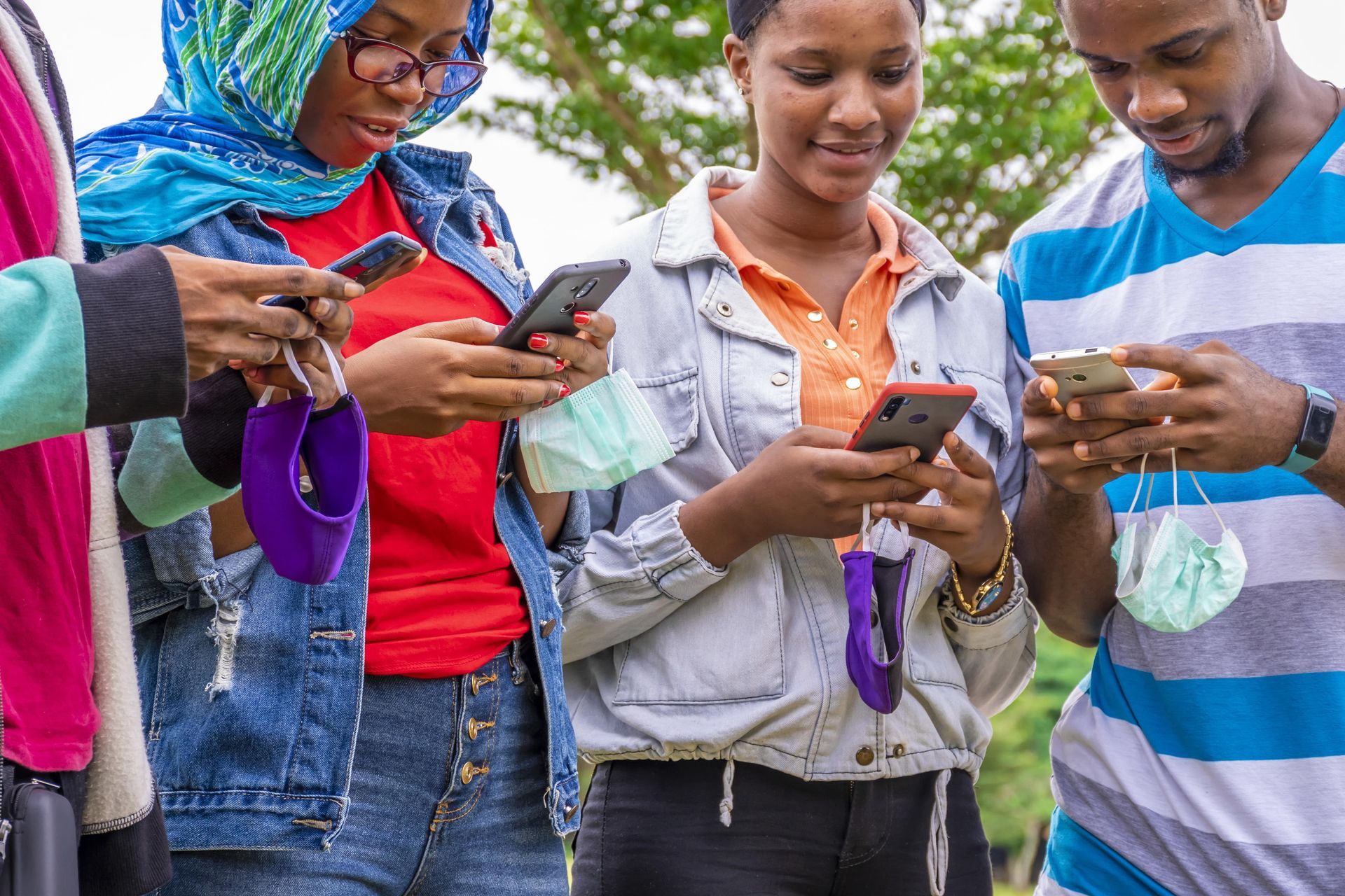 Quatre individus à l'extérieur, chacun concentré sur son propre smartphone, tenant des masques faciaux à la main.