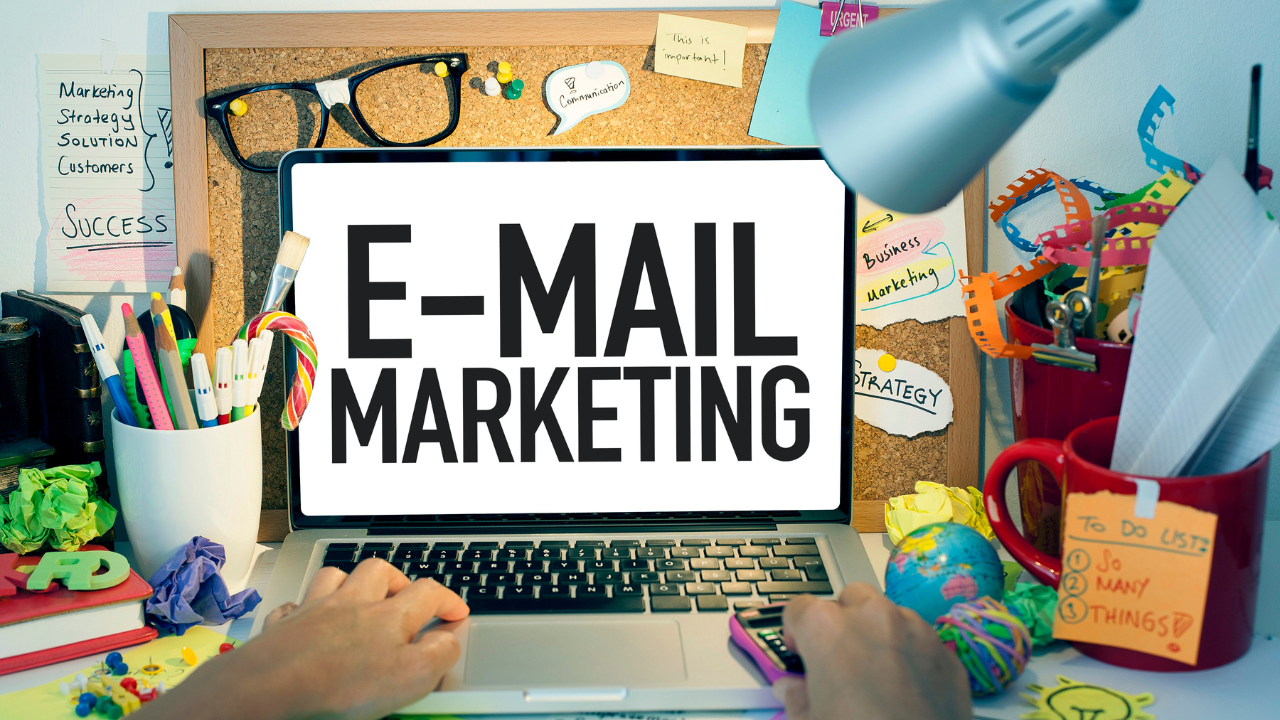 Une personne tape sur un ordinateur portable affichant le texte « E-MAIL MARKETING » sur un bureau encombré.