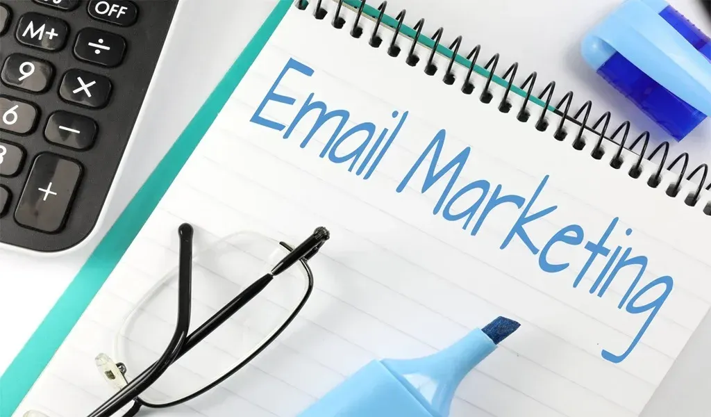 Un carnet avec l'inscription « Marketing par e-mail » sur la page, à côté d'une calculatrice, de lunettes et de surligneurs bleus.