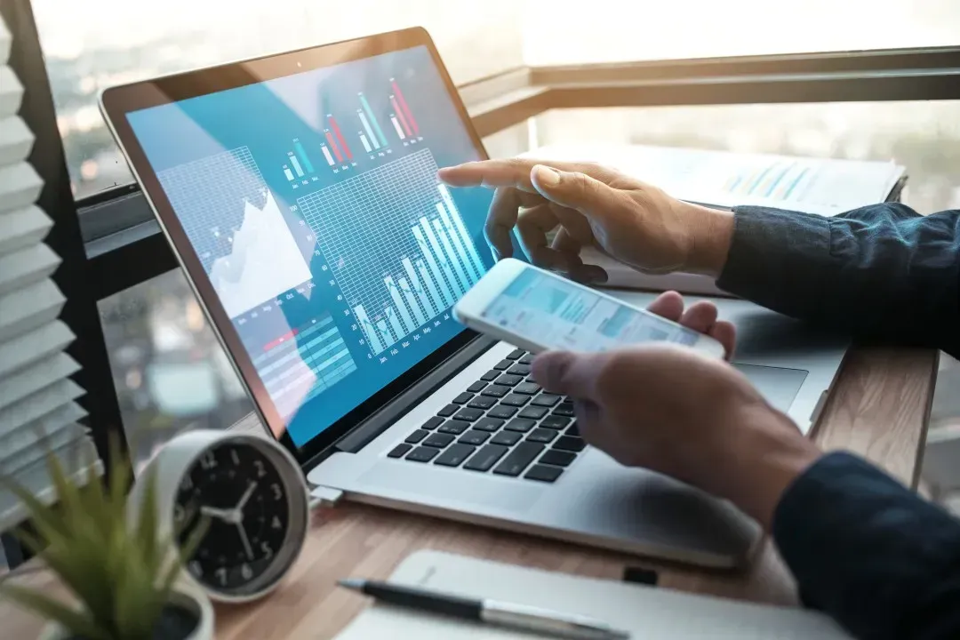 Une personne travaillant à un bureau, pointant du doigt des graphiques de données financières sur l'écran d'un ordinateur portable tout en tenant un smartphone.