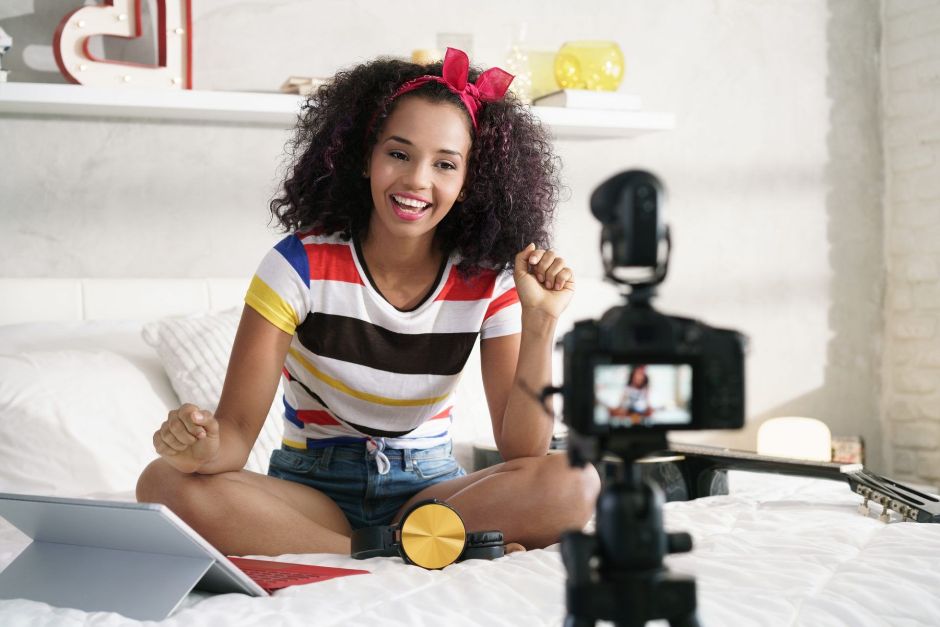 Une personne souriante, aux cheveux bouclés et portant un bandeau rouge, enregistre une vidéo assise sur un lit avec une caméra et une tablette.