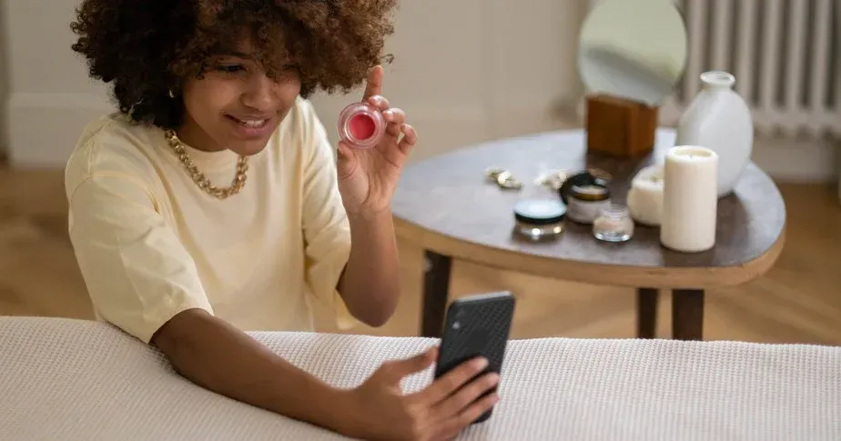 Une personne aux cheveux bouclés sourit en tenant un petit pot de crème rose et en regardant l'écran d'un smartphone.