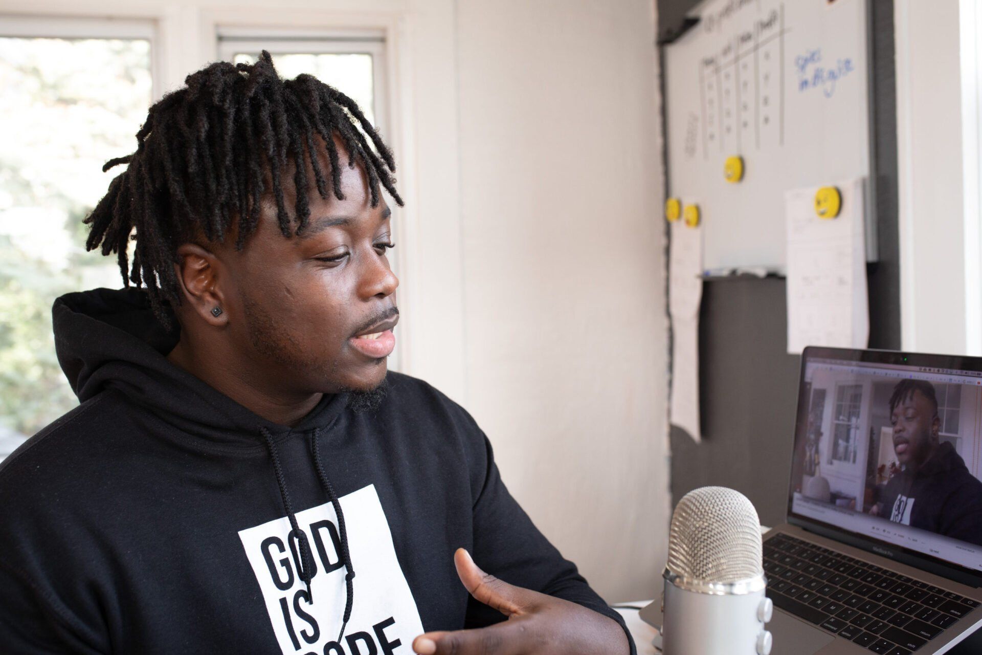 Une personne portant un sweat-shirt à capuche noir avec du texte parle dans un microphone tout en regardant son reflet sur l'écran d'un ordinateur portable.