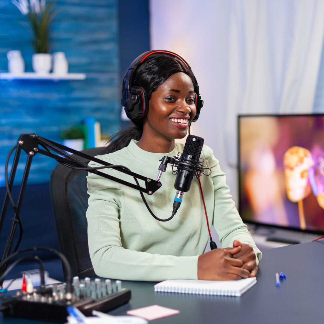Une personne portant un casque audio parle dans un microphone à un bureau, souriante, un carnet et une table de mixage audio devant elle.