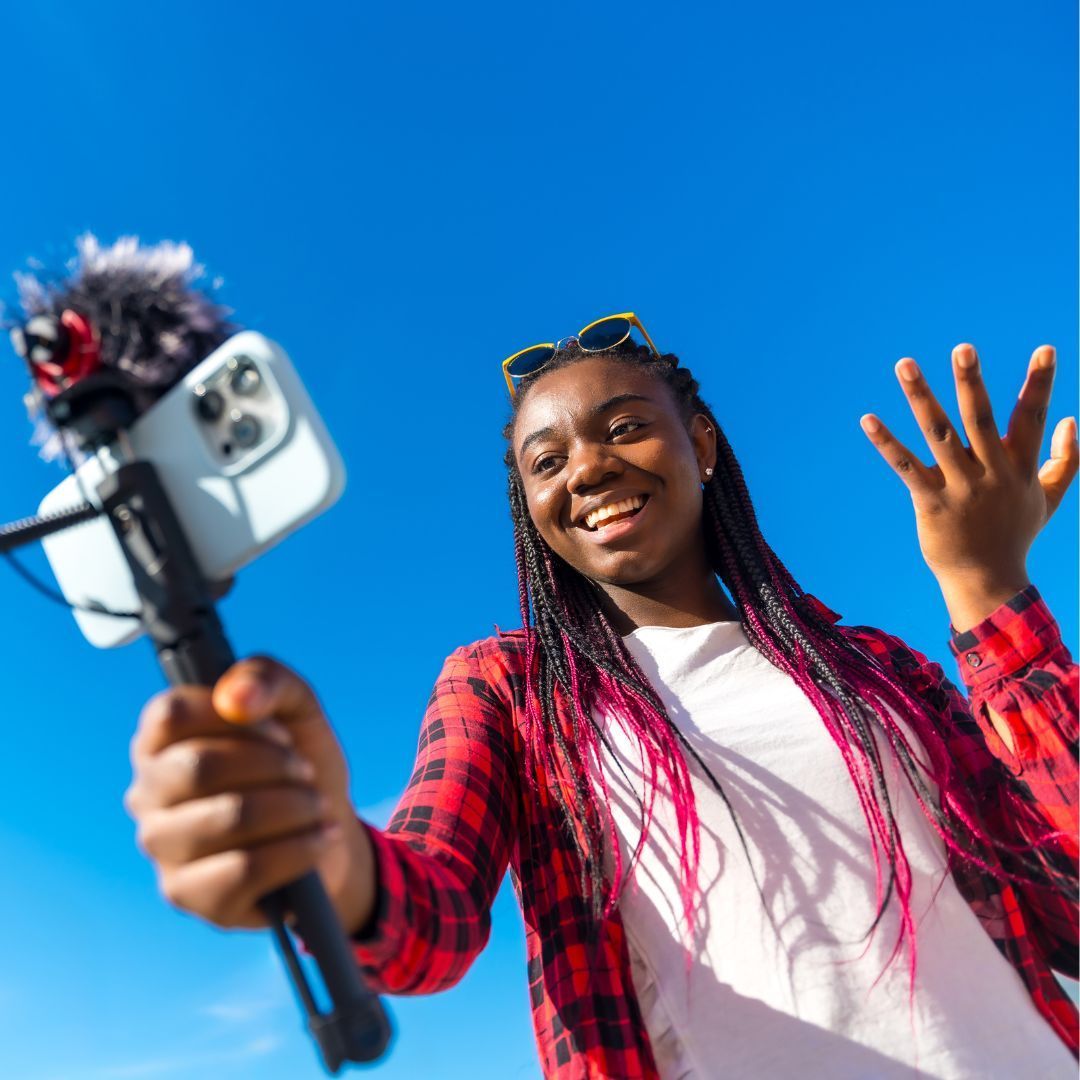 Une personne tenant un téléphone sur un trépied avec un microphone, se détachant sur un ciel bleu éclatant, souriant et gesticulant tout en enregistrant.