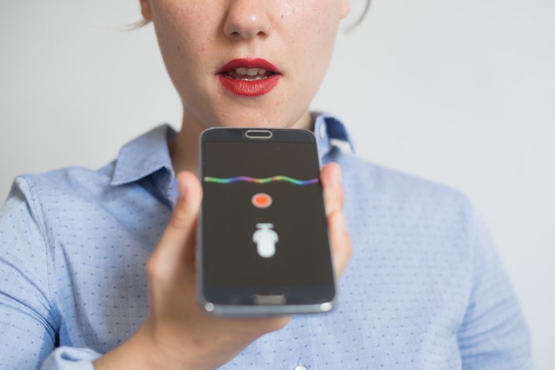 Une personne vêtue d'une chemise bleu clair tient un smartphone affichant l'interface d'une application d'enregistrement avec une onde sonore et une icône.