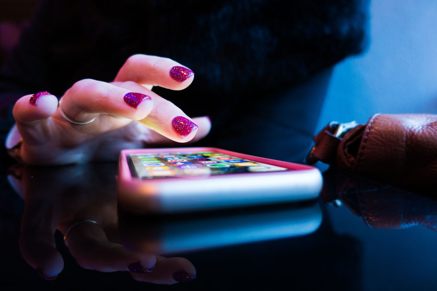 Une personne aux ongles vernis utilise son index pour toucher l'écran d'un smartphone posé sur une surface réfléchissante.
