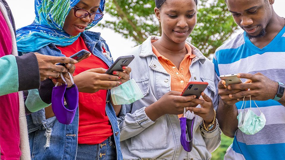 Quatre personnes, les yeux rivés sur leur smartphone, chacune tenant un masque, en extérieur dans un cadre décontracté.