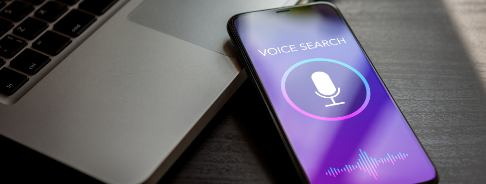 Un smartphone affichant une interface de recherche vocale avec une icône de microphone sur un écran violet, à côté d'un clavier d'ordinateur portable.
