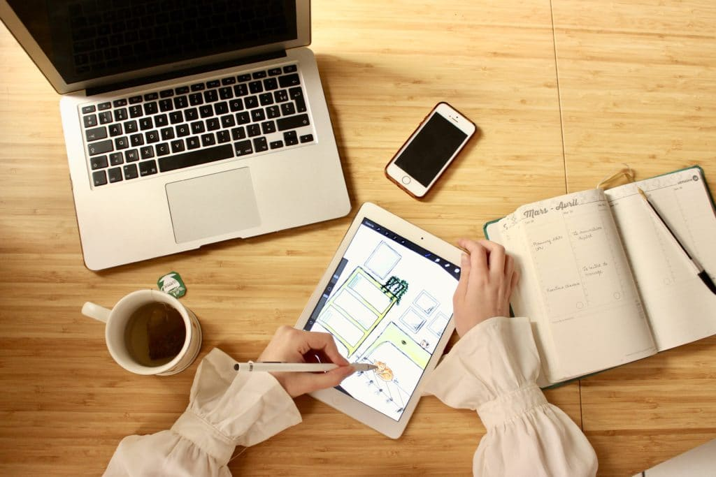 Une personne utilise un stylet numérique sur une tablette posée à côté d'un ordinateur portable, d'un smartphone, d'un carnet ouvert et d'une tasse sur un bureau en bois.