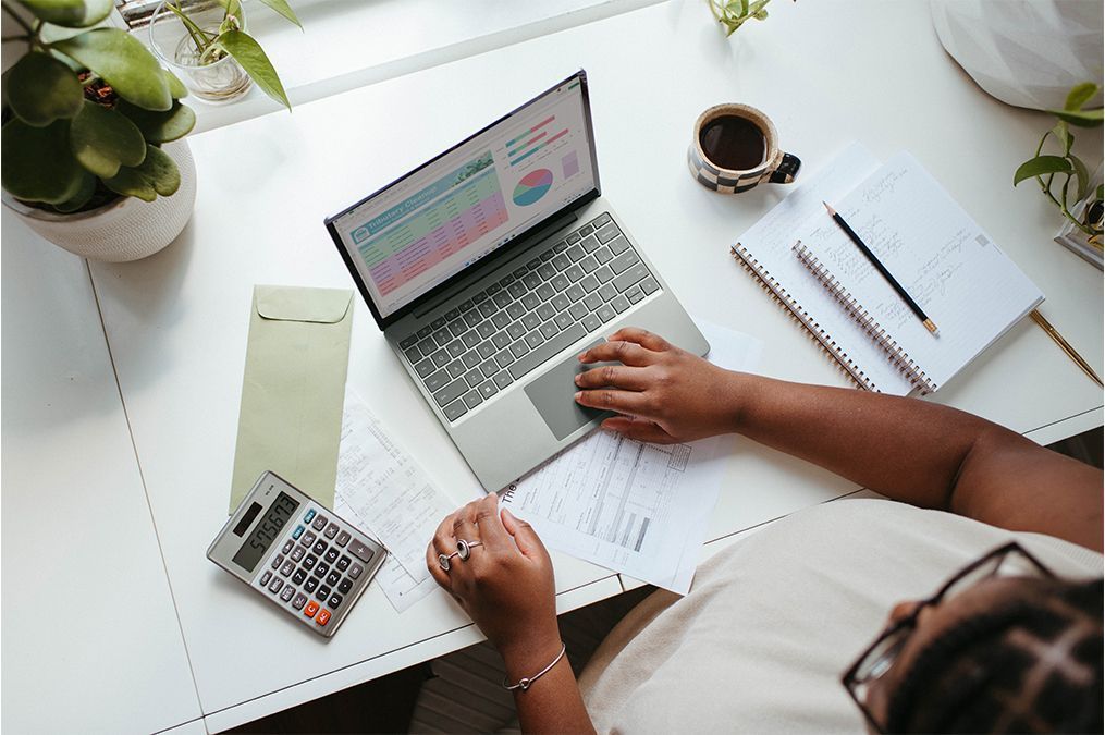 Une personne travaille à un bureau blanc avec un ordinateur portable affichant des graphiques, une calculatrice, un carnet et une plante en pot.