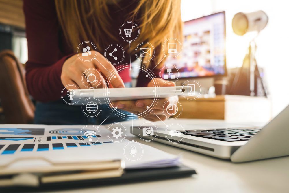 Une personne tenant une tablette au-dessus de documents sur un bureau, avec des icônes numériques représentant le monde des affaires et la connectivité internet.