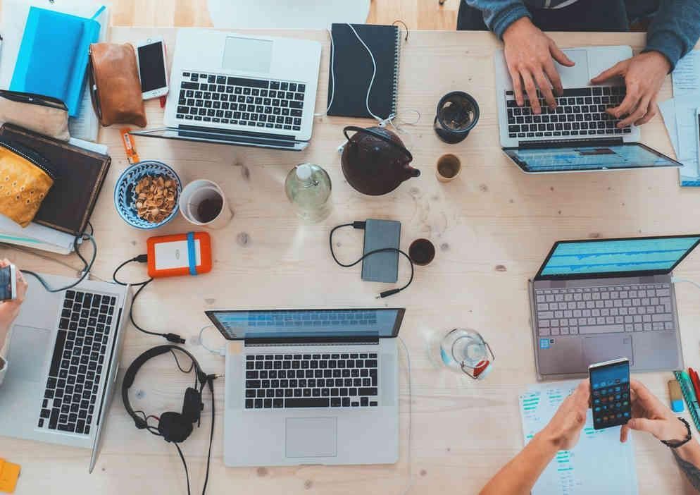 Vue de dessus d'une table en bois clair sur laquelle sont posés plusieurs ordinateurs portables, appareils mobiles, casques audio et carnets.