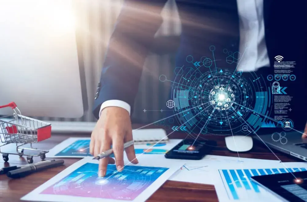 Une personne en costume analyse des documents commerciaux et une interface de visualisation de données numériques sur un bureau où se trouve un chariot de courses.
