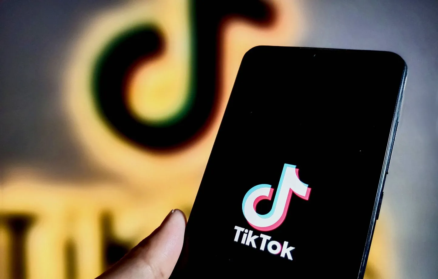 Une main tient un smartphone affichant le logo de l'application TikTok, avec un grand logo TikTok flou en arrière-plan.
