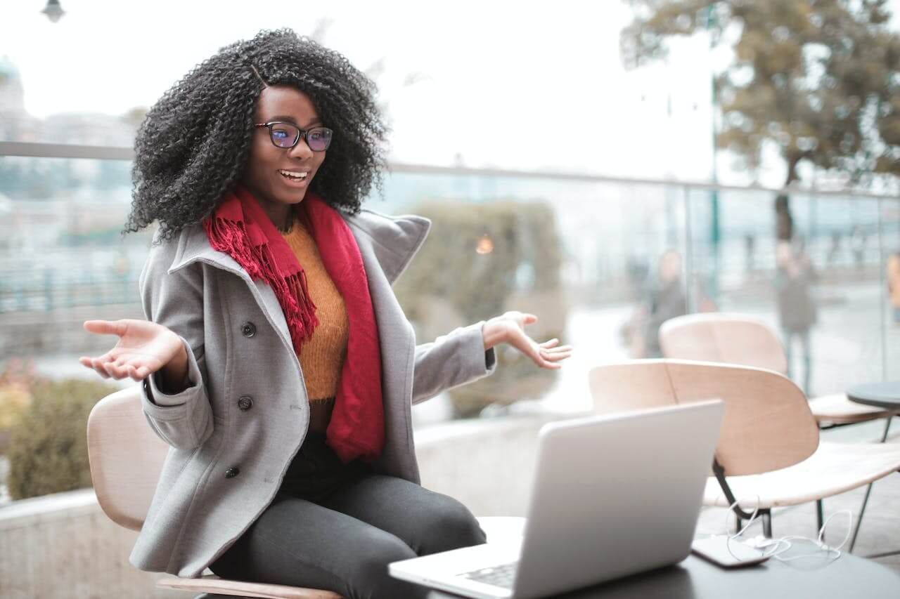 Une personne portant un manteau gris et une écharpe rouge, assise à une table avec un ordinateur portable, gesticulant en souriant.