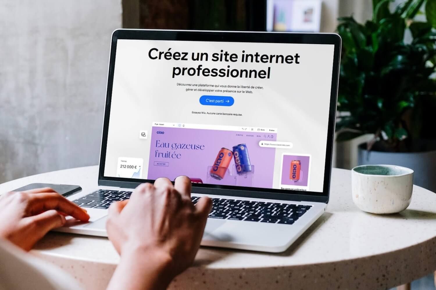 Une personne travaille sur un ordinateur portable, utilisant un logiciel professionnel de création de sites web, avec une tasse de café et une plante sur une table ronde.