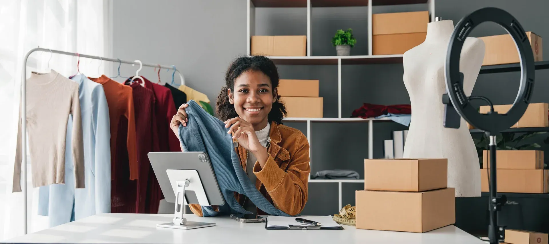 Un entrepreneur souriant est assis à un bureau dans un atelier de confection, tenant un pull gris devant une tablette et un anneau lumineux.