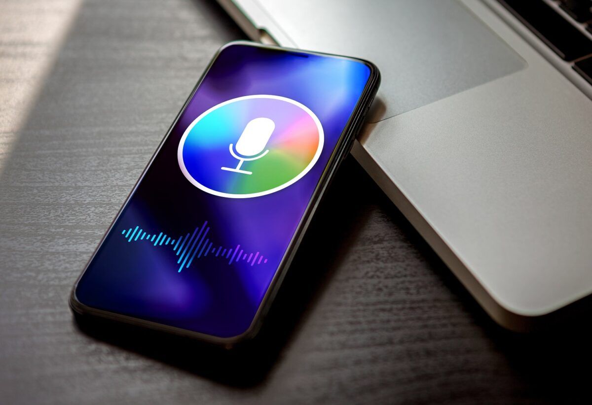 Un smartphone affichant l'interface de l'assistant vocal Siri sur son écran, posé sur un bureau sombre à côté d'un ordinateur portable.