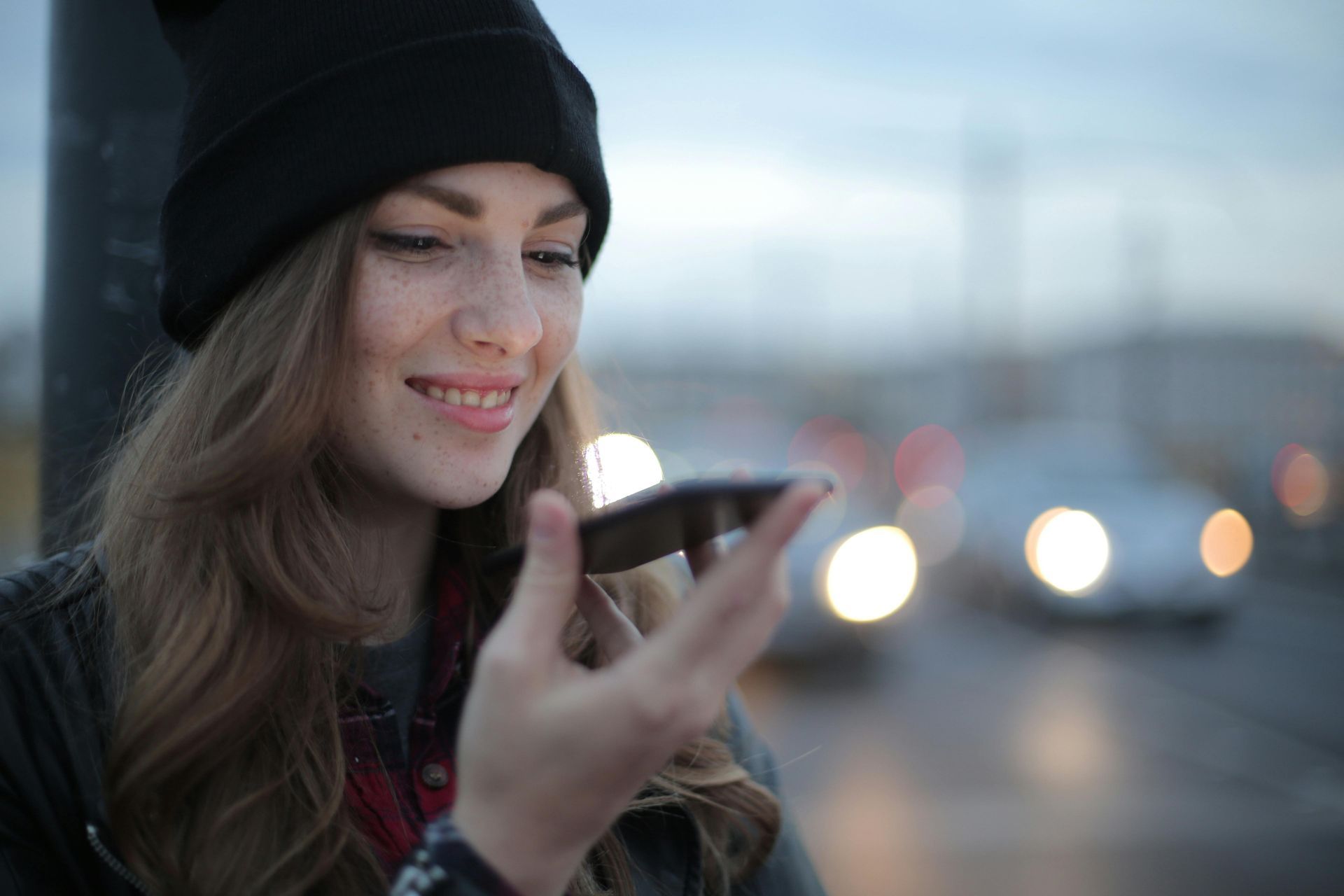 Une personne portant un bonnet noir sourit en parlant dans un smartphone tenu près de son visage, sur fond de paysage urbain flou.