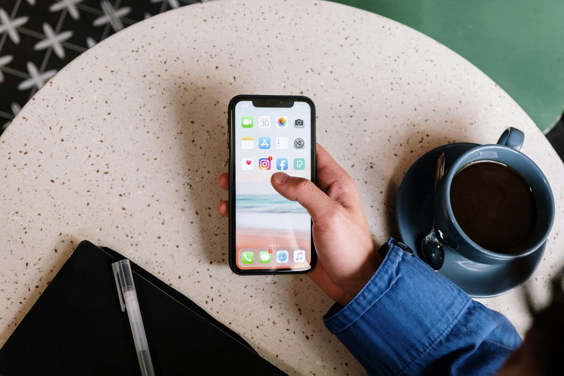 Une main tient un smartphone avec un fond d'écran de plage, vu de dessus sur une table mouchetée à côté d'un café et d'un stylo.