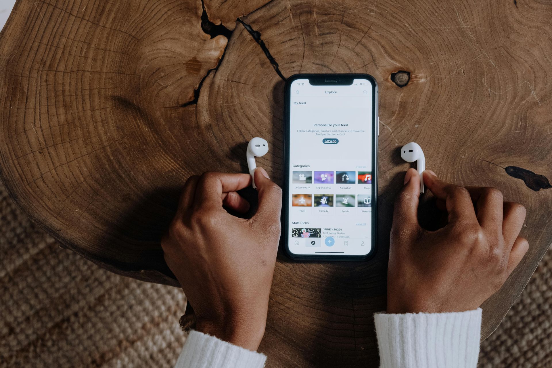 Des mains tenant des écouteurs sans fil blancs près d'un smartphone affichant une application musicale, posé sur une table en bois rustique.