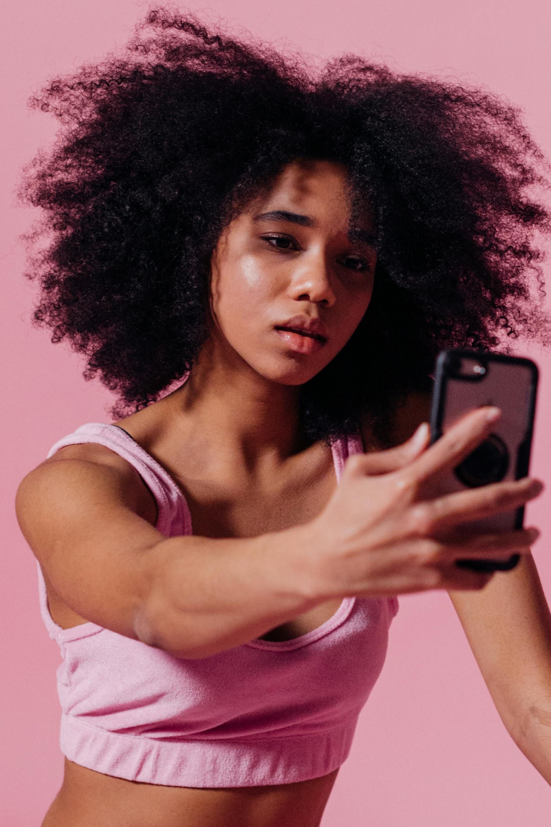 Une personne aux cheveux bouclés, foncés et volumineux, vêtue d'un haut rose, tient un smartphone pour prendre un selfie devant un fond rose.