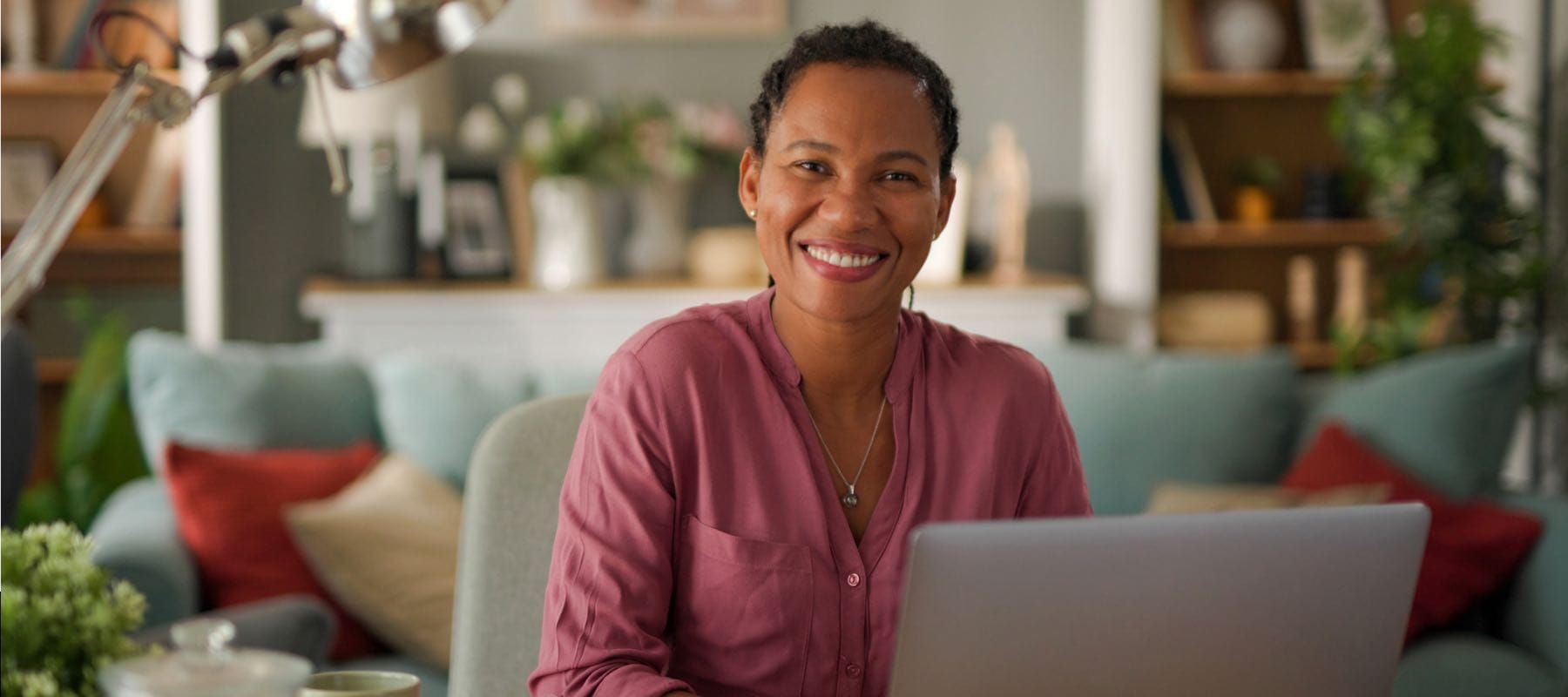 Une personne vêtue d'une chemise magenta est assise à un bureau avec un ordinateur portable, souriant dans un décor de bureau à domicile flou.