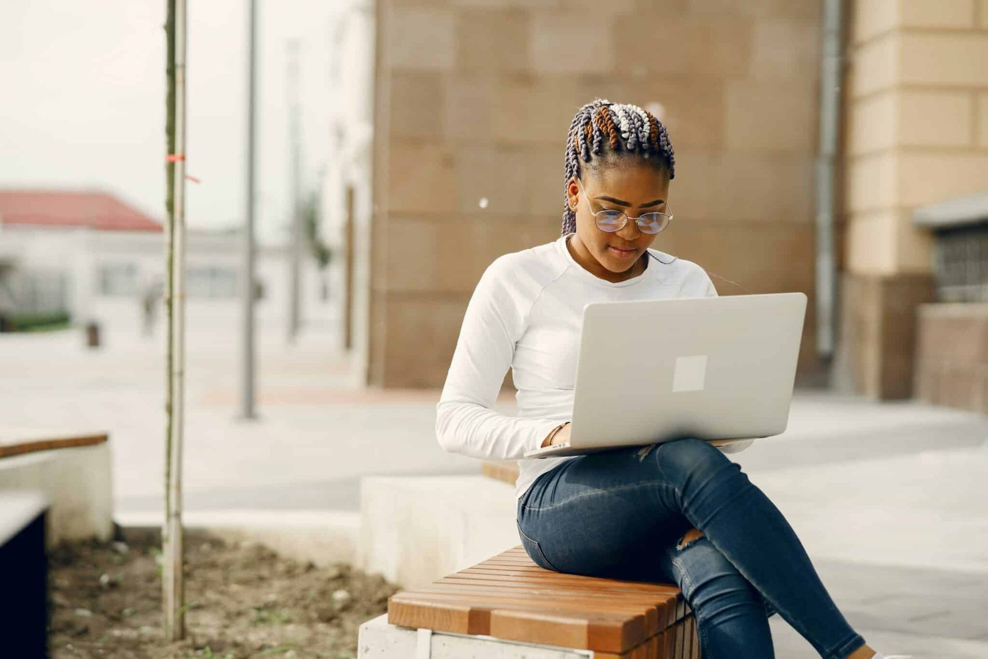 Une personne avec des tresses et des lunettes est assise sur un banc à l'extérieur, en train de taper sur un ordinateur portable.