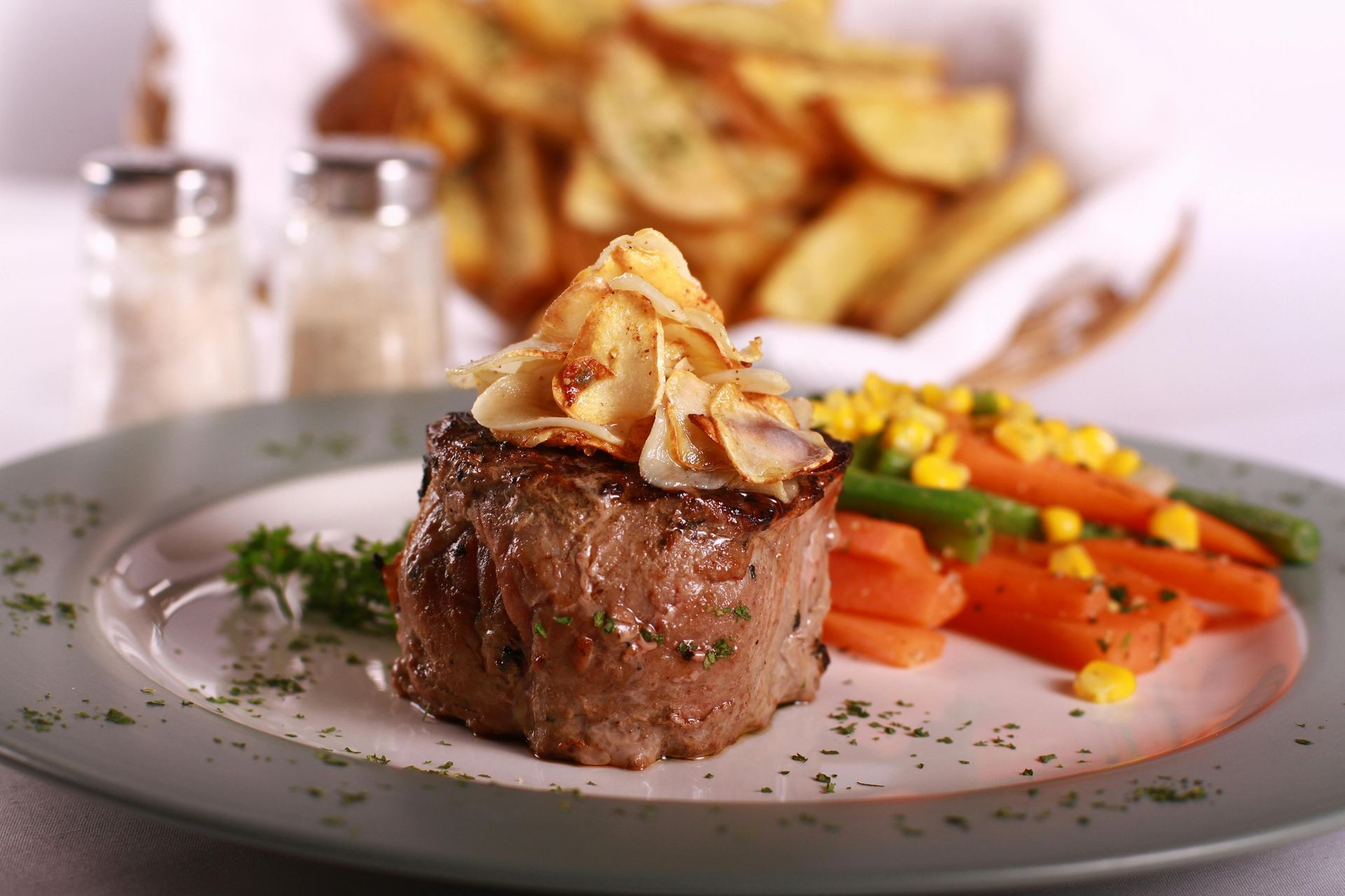 Steak medallion topped with garnish, served with carrots and fries on a white plate, restaurant-style plating