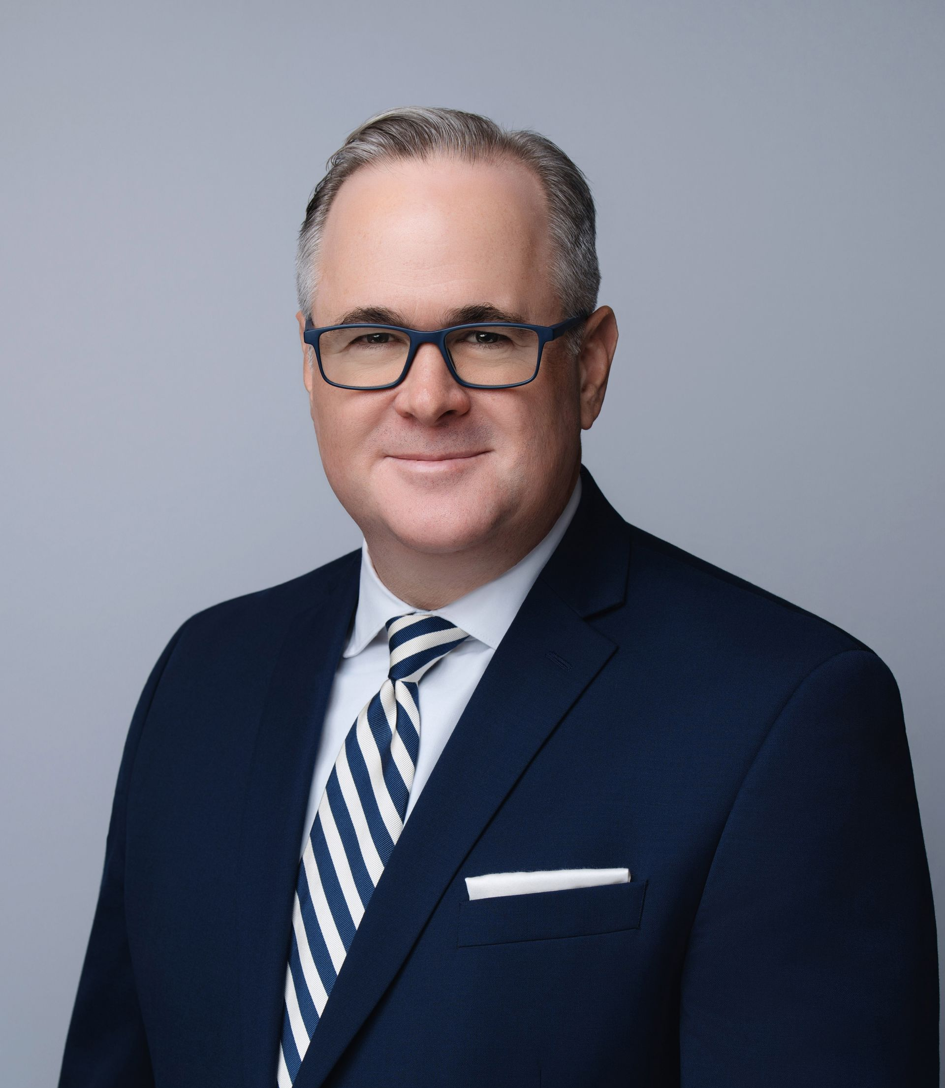 Man in a navy suit and glasses, smiling, with a striped tie, posing against a gray backdrop.