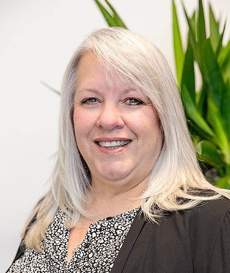 A woman with blonde hair is smiling in front of a plant.