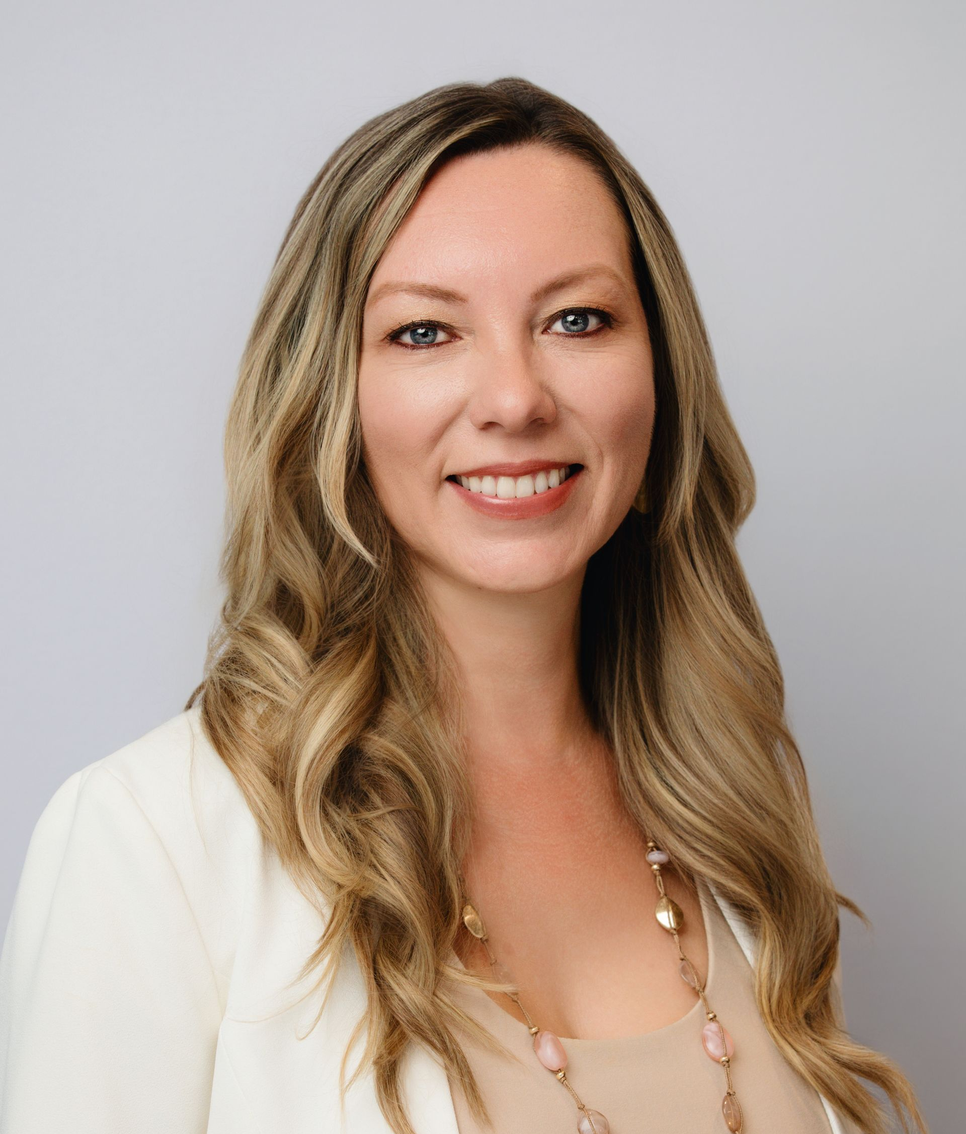 Woman with long blonde hair smiles, wearing a cream blazer and necklace, against a gray background.