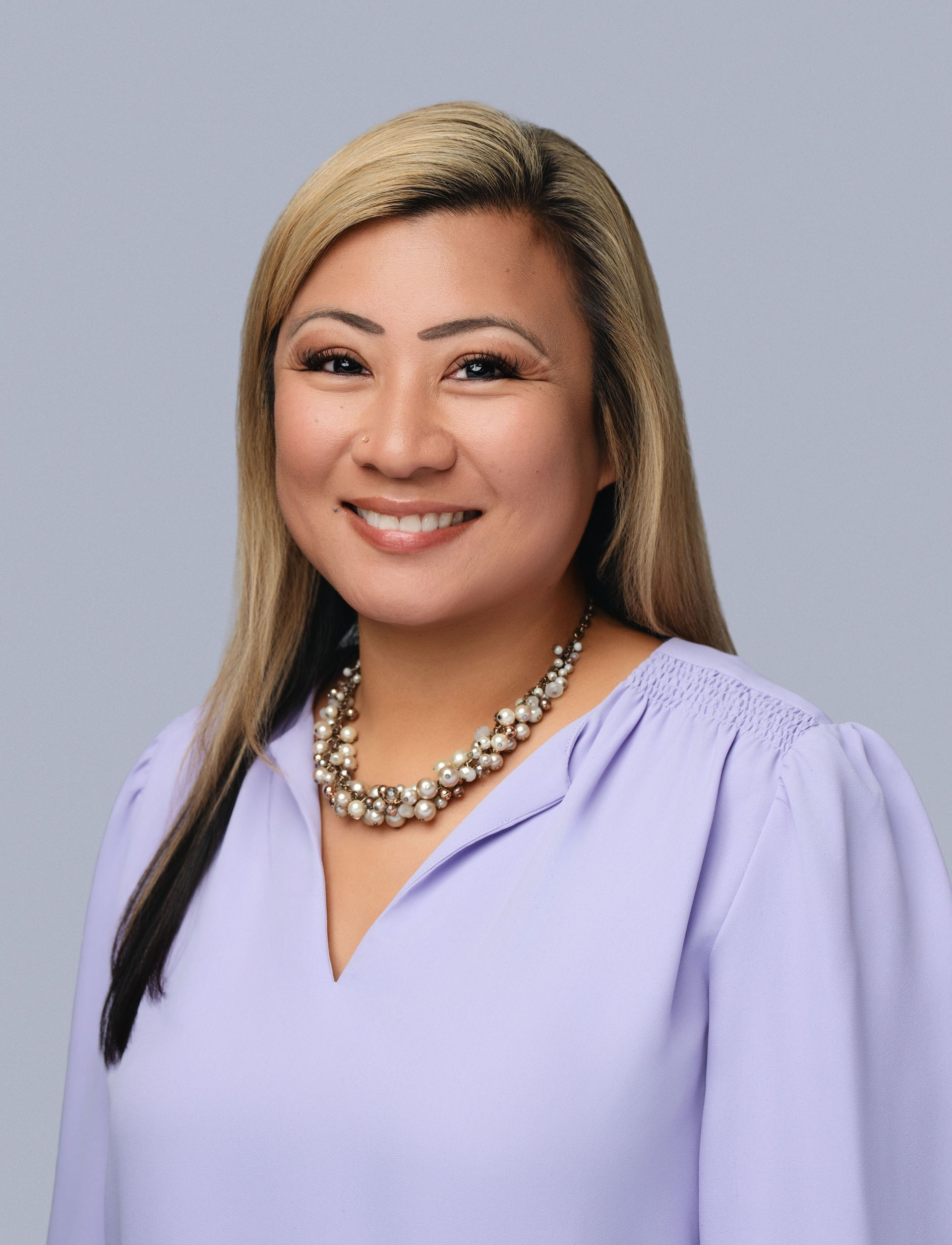 Smiling professional in a lavender blouse and necklace against a gray studio background