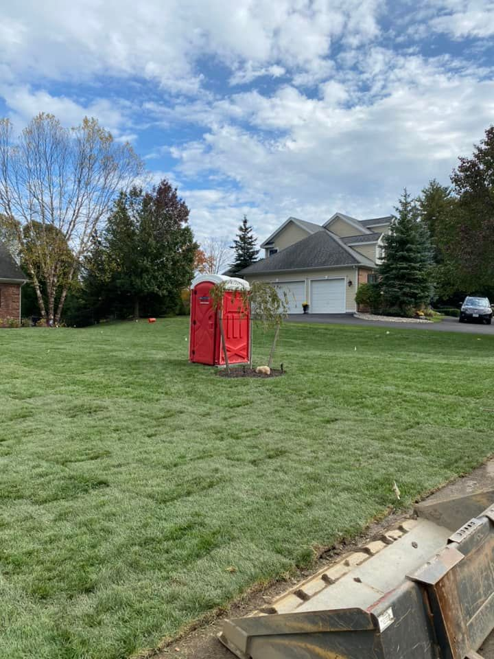 Red portable toilet on a green lawn in front of a house, under a cloudy sky.