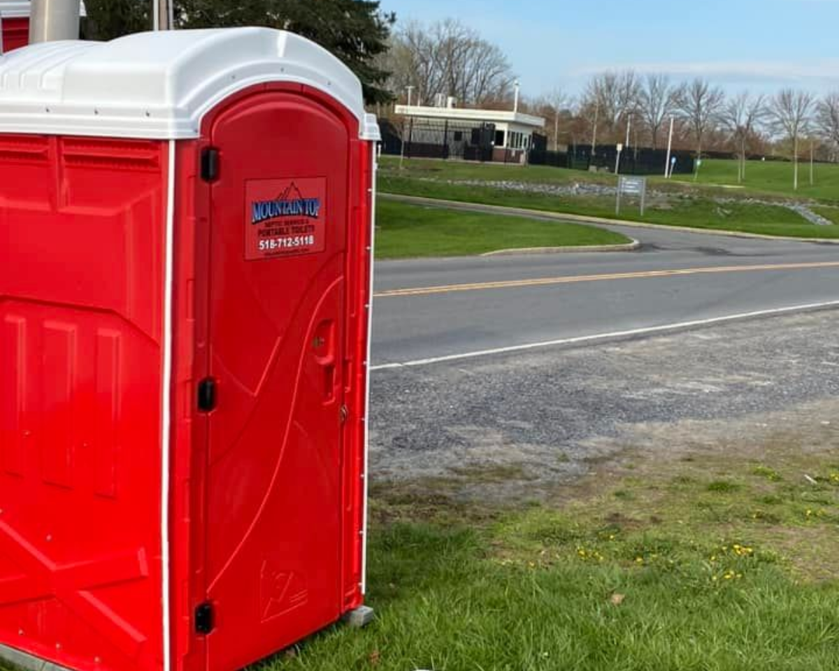 Red portable toilet on green grass next to a road, with a blue sky in the background.