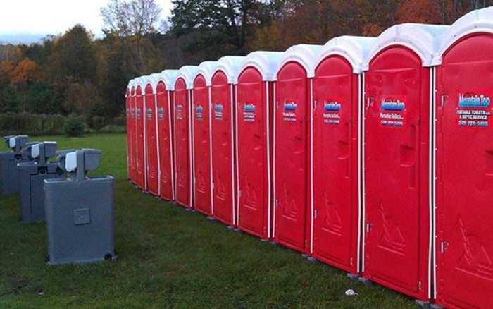 Row of red portable toilets on grass with trees in the background.