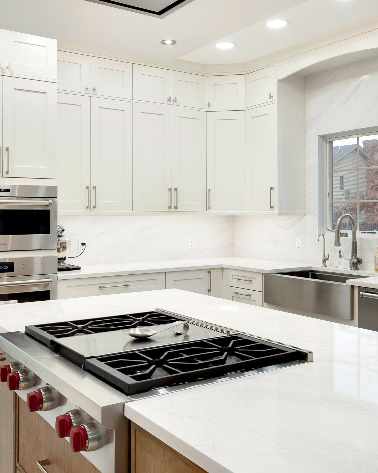 Bright white modern kitchen with large island, gas cooktop, built-in ovens, and sink by a window