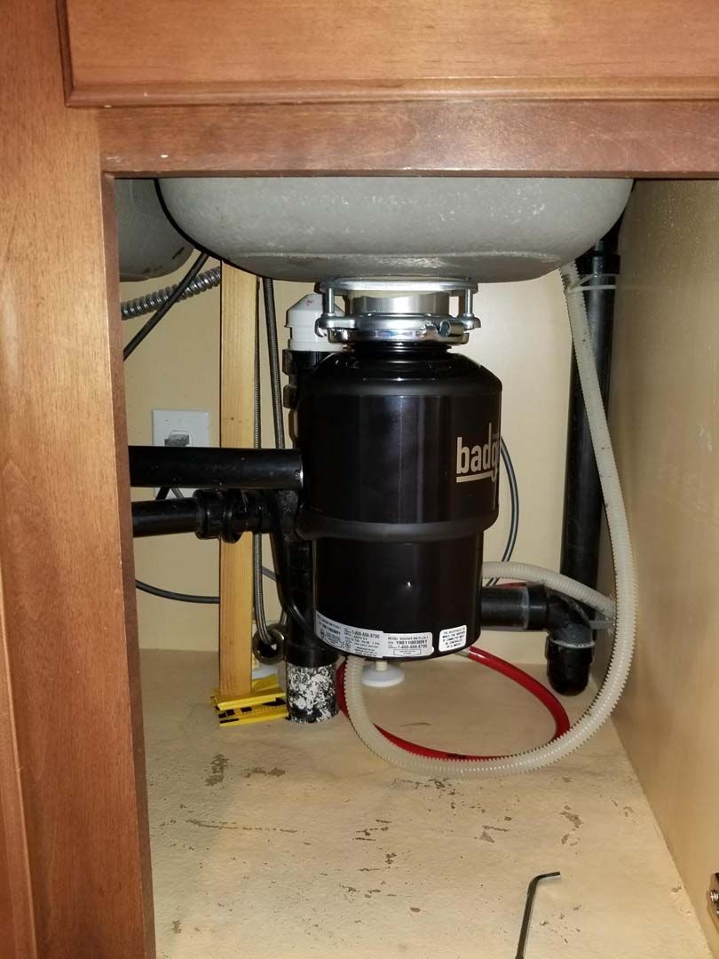 Under-sink view: A black garbage disposal is attached to the kitchen sink pipes. Light-colored cabinet.
